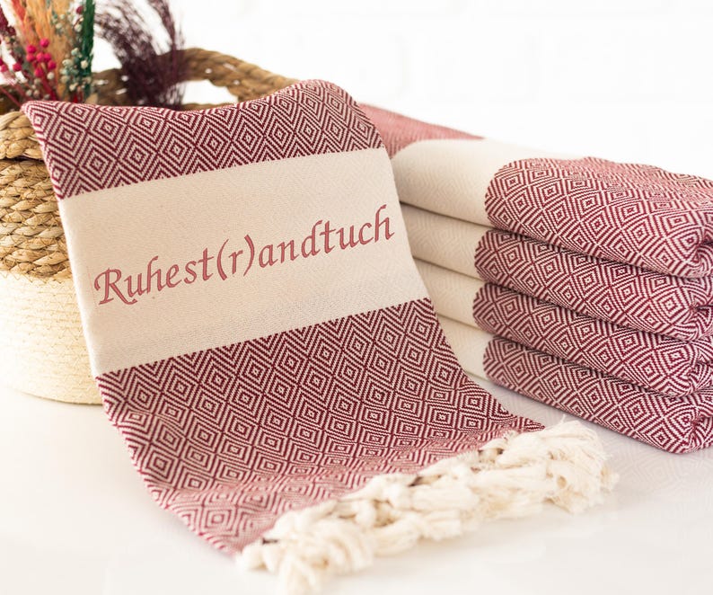 Puede incluir: Toallas burdeos y crema plegadas con un patr&oacute;n geom&eacute;trico de diamantes. Una toalla est&aacute; drapeada hacia adelante, mostrando el texto "Ruhest(r)andtuch" en burdeos. Las toallas tienen flecos color crema.