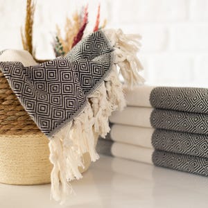 Puede incluir: Una pila de toallas blancas y grises plegadas con estampado de diamantes y bordes con flecos. Una toalla a juego est&aacute; sobre una cesta tejida. Plantas decorativas secas en el fondo. Las toallas son de material suave.