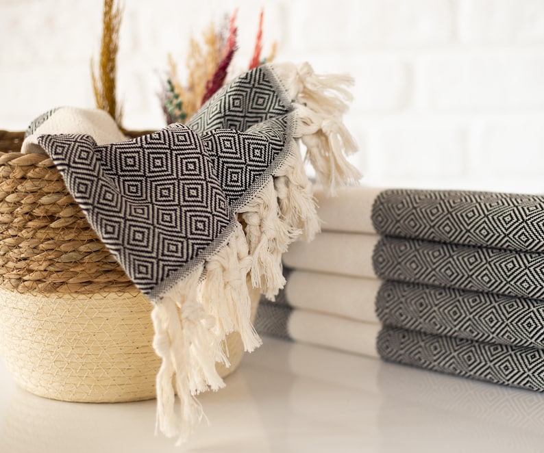 Puede incluir: Una pila de toallas blancas y grises plegadas con estampado de diamantes y bordes con flecos. Una toalla a juego est&aacute; sobre una cesta tejida. Plantas decorativas secas en el fondo. Las toallas son de material suave.