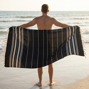 Puede incluir: Una toalla de playa a rayas blancas y negras con bordes con flecos es sostenida abierta por una persona en una playa de arena. La toalla tiene rayas verticales y est&aacute; hecha de un material suave y ligero. El oc&eacute;ano es visible en el fondo.