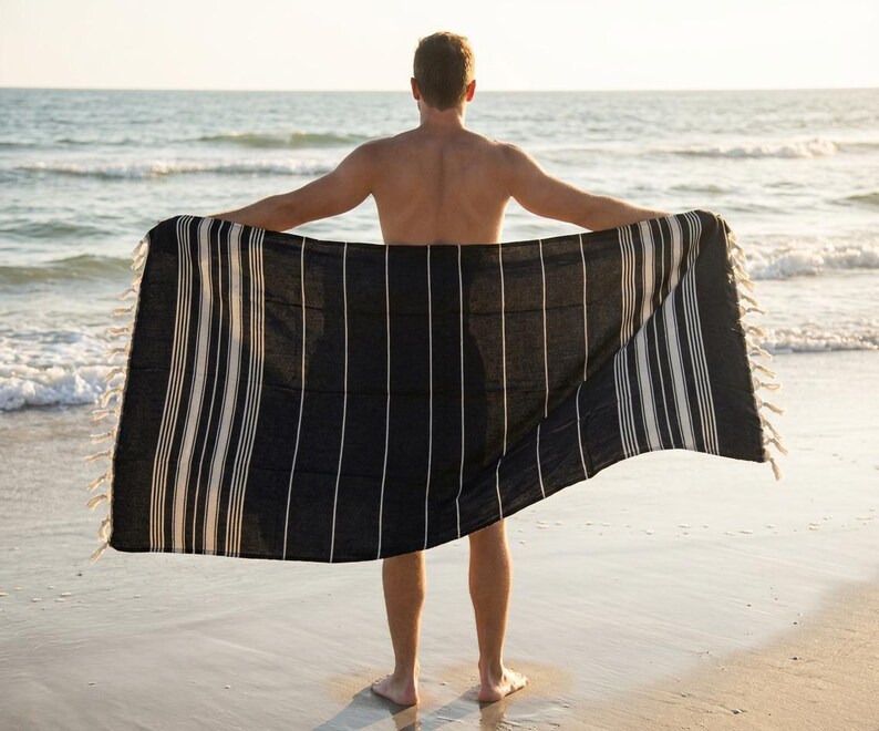 Puede incluir: Una toalla de playa a rayas blancas y negras con bordes con flecos es sostenida abierta por una persona en una playa de arena. La toalla tiene rayas verticales y est&aacute; hecha de un material suave y ligero. El oc&eacute;ano es visible en el fondo.