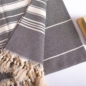 Puede incluir: Una toalla doblada a rayas grises y blancas con flecos color crema. La toalla tiene un tejido texturizado y un dise&ntilde;o sencillo y cl&aacute;sico. Se ve un peque&ntilde;o jabonera de madera con una pastilla de jab&oacute;n.