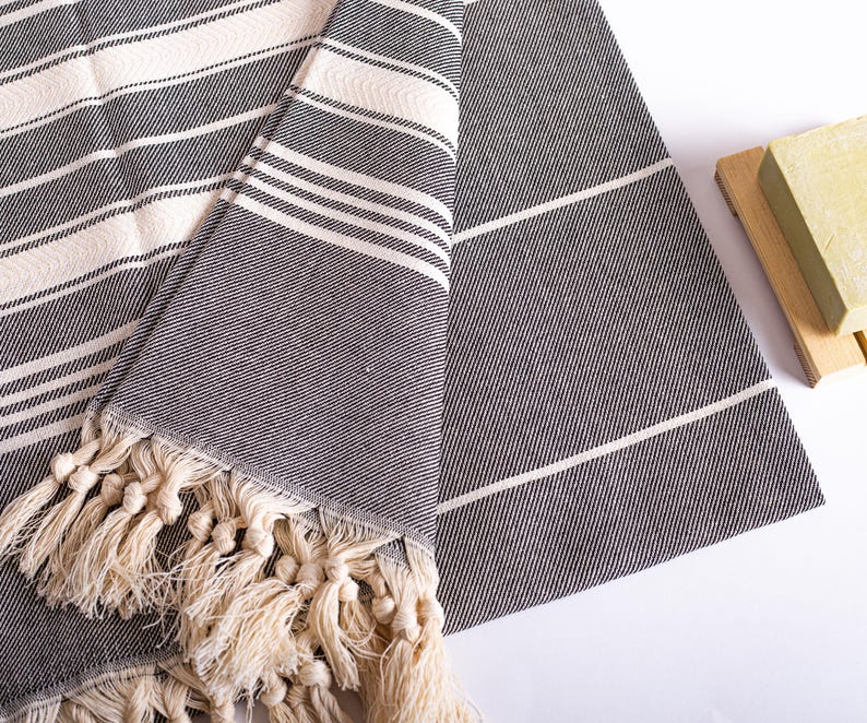 Puede incluir: Una toalla doblada a rayas grises y blancas con flecos color crema. La toalla tiene un tejido texturizado y un dise&ntilde;o sencillo y cl&aacute;sico. Se ve un peque&ntilde;o jabonera de madera con una pastilla de jab&oacute;n.