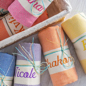 Puede incluir: Toallas enrolladas con estampado de diamantes en varios colores, como naranja, rosa, azul, morado y amarillo. Cada toalla est&aacute; envuelta con una banda blanca bordada con un nombre y atada con hilo. Algunas est&aacute;n en una cesta.