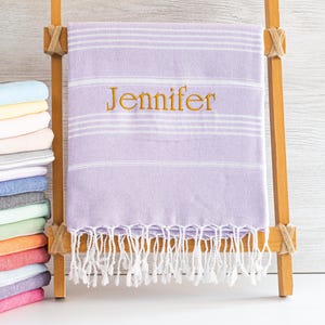 May include: A lavender and white striped Turkish towel with the name "Jennifer" embroidered in gold. The towel has a fringe and is displayed on a wooden ladder. A stack of colorful towels is visible on the left.