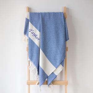 Puede incluir: Toalla azul y blanca estampada con el nombre "Taylor" bordado en azul. La toalla est&aacute; colgada de una escalera de madera. La toalla tiene un dise&ntilde;o geom&eacute;trico y flecos blancos.