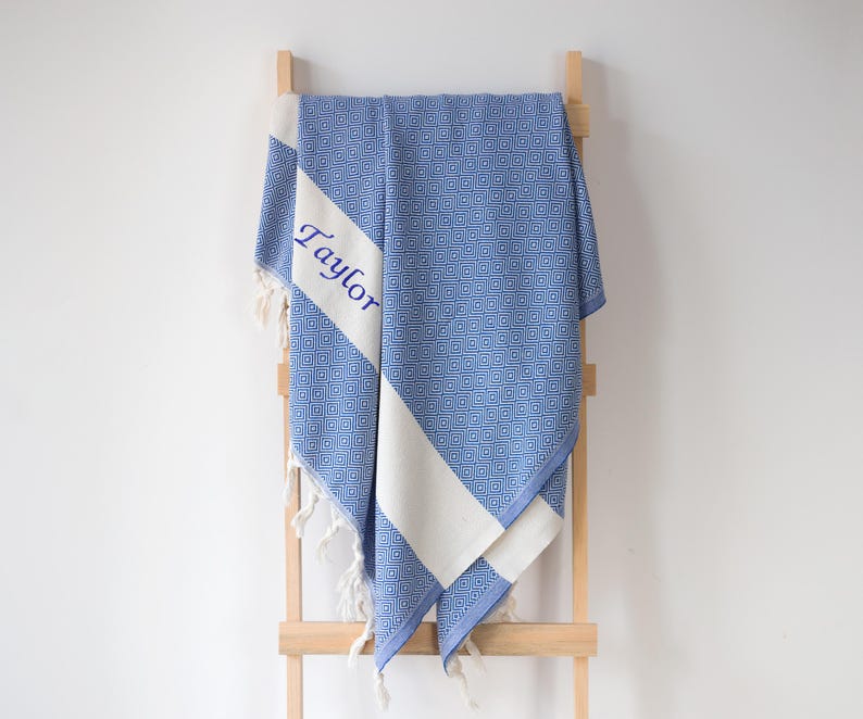 Puede incluir: Toalla azul y blanca estampada con el nombre "Taylor" bordado en azul. La toalla est&aacute; colgada de una escalera de madera. La toalla tiene un dise&ntilde;o geom&eacute;trico y flecos blancos.