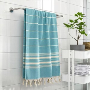 Puede incluir: Una toalla a rayas turquesa y blanca con flecos cuelga de un toallero cromado. La toalla est&aacute; en un ba&ntilde;o con azulejos blancos. Una peque&ntilde;a planta en maceta y toallas est&aacute;n en un estante blanco.
