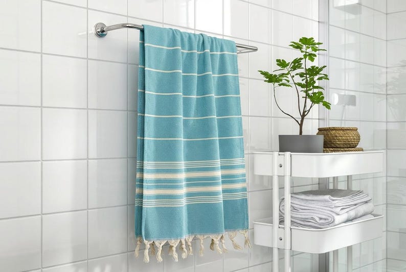Puede incluir: Una toalla a rayas turquesa y blanca con flecos cuelga de un toallero cromado. La toalla est&aacute; en un ba&ntilde;o con azulejos blancos. Una peque&ntilde;a planta en maceta y toallas est&aacute;n en un estante blanco.