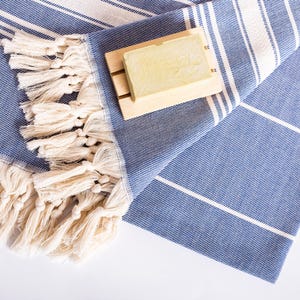 Puede incluir: Una toalla turca a rayas azules y blancas con flecos color crema. Una jabonera de madera sostiene una pastilla de jab&oacute;n. La toalla est&aacute; doblada y tiene un dise&ntilde;o sencillo y limpio, ideal para un ba&ntilde;o o la playa.