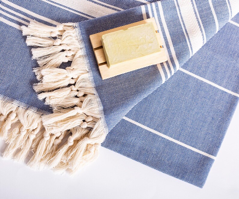 Puede incluir: Una toalla turca a rayas azules y blancas con flecos color crema. Una jabonera de madera sostiene una pastilla de jab&oacute;n. La toalla est&aacute; doblada y tiene un dise&ntilde;o sencillo y limpio, ideal para un ba&ntilde;o o la playa.