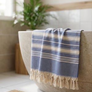 Puede incluir: Una toalla a rayas azules y blancas con flecos cuelga del borde de una ba&ntilde;era de piedra. La toalla tiene rayas horizontales y un aspecto texturizado. El entorno del ba&ntilde;o incluye una planta y luz natural.