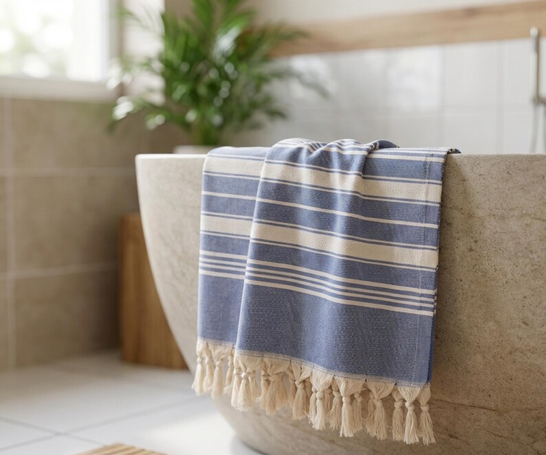 Puede incluir: Una toalla a rayas azules y blancas con flecos cuelga del borde de una ba&ntilde;era de piedra. La toalla tiene rayas horizontales y un aspecto texturizado. El entorno del ba&ntilde;o incluye una planta y luz natural.