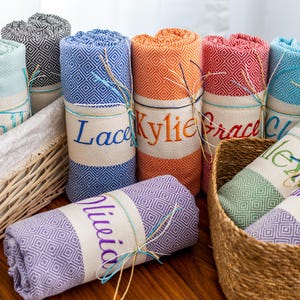 Puede incluir: Toallas enrolladas y personalizadas en varios colores, incluyendo azul, naranja y rojo, con patrones de diamantes. Cada toalla est&aacute; envuelta con una banda que muestra un nombre en letras bordadas. Algunas toallas est&aacute;n en cestas de mimbre.