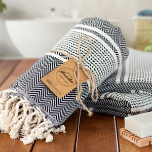 May include: Rolled black and white patterned towel with a tag tied with twine. The towel has a geometric design and fringe. A second folded towel and a bar of soap on a wooden soap dish are also visible.