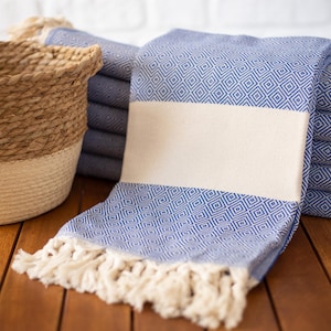 Puede incluir: Toallas azules y blancas con estampado de diamantes, con una franja crema y flecos. Una cesta tejida con tonos naturales y crema est&aacute; en el fondo. Las toallas est&aacute;n apiladas sobre una superficie de madera.