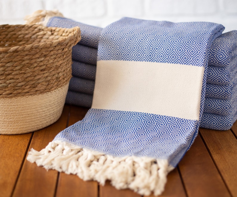 Puede incluir: Toallas azules y blancas con estampado de diamantes, con una franja crema y flecos. Una cesta tejida con tonos naturales y crema est&aacute; en el fondo. Las toallas est&aacute;n apiladas sobre una superficie de madera.