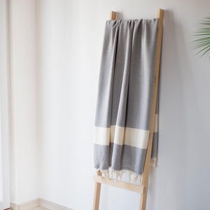 Puede incluir: Una manta gris y blanca estampada con rayas y borlas color crema est&aacute; colgada sobre una escalera de madera clara. La manta cuelga contra una pared blanca, con una ventana y vegetaci&oacute;n en el fondo. Mide aproximadamente 180 cm de largo.