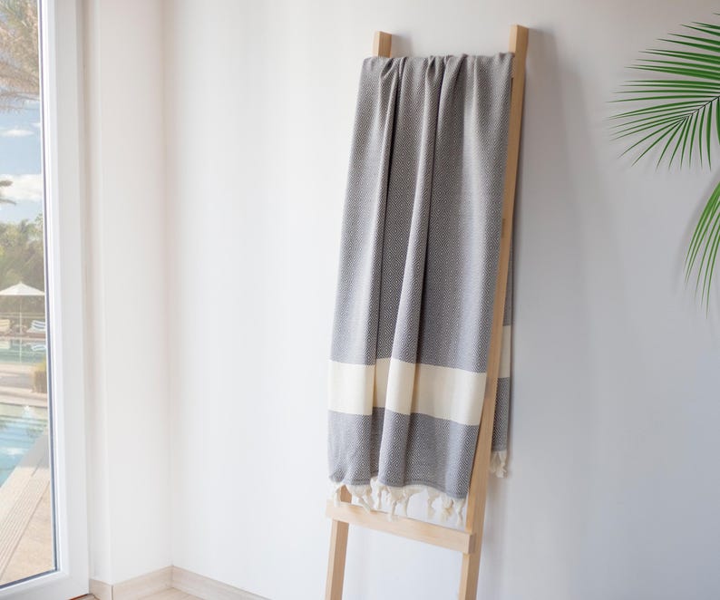 Puede incluir: Una manta gris y blanca estampada con rayas y borlas color crema est&aacute; colgada sobre una escalera de madera clara. La manta cuelga contra una pared blanca, con una ventana y vegetaci&oacute;n en el fondo. Mide aproximadamente 180 cm de largo.