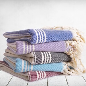 Puede incluir: Una pila de toallas plegadas a rayas en varios colores: azul, lavanda, gris y rojo. Cada toalla tiene rayas blancas y un borde con flecos. Una manta crema con flecos est&aacute; drapeada sobre la pila.