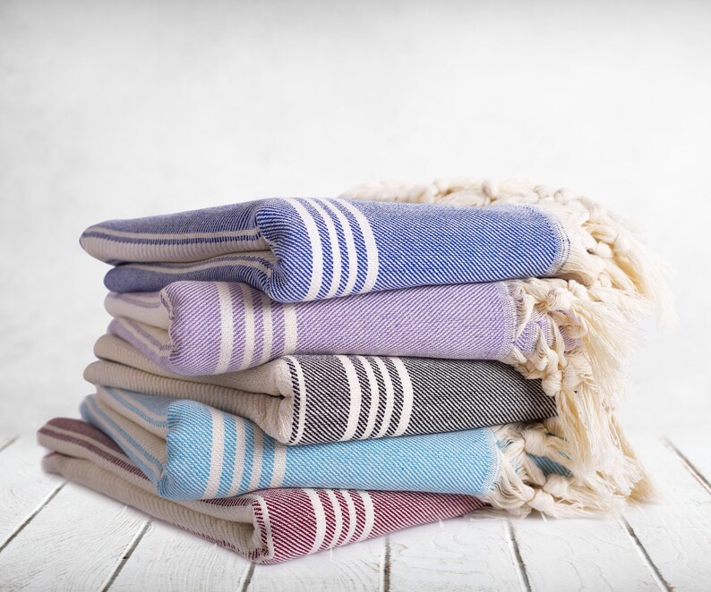 Puede incluir: Una pila de toallas plegadas a rayas en varios colores: azul, lavanda, gris y rojo. Cada toalla tiene rayas blancas y un borde con flecos. Una manta crema con flecos est&aacute; drapeada sobre la pila.