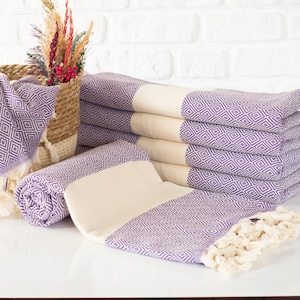 Puede incluir: Una pila de toallas moradas y crema con estampado de diamantes y bordes con flecos. Tambi&eacute;n se ven una toalla enrollada y una cesta con flores decorativas. Las toallas est&aacute;n dispuestas sobre una superficie blanca contra una pared de ladrillos blancos.