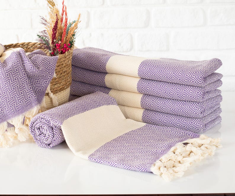 Puede incluir: Una pila de toallas moradas y crema con estampado de diamantes y bordes con flecos. Tambi&eacute;n se ven una toalla enrollada y una cesta con flores decorativas. Las toallas est&aacute;n dispuestas sobre una superficie blanca contra una pared de ladrillos blancos.