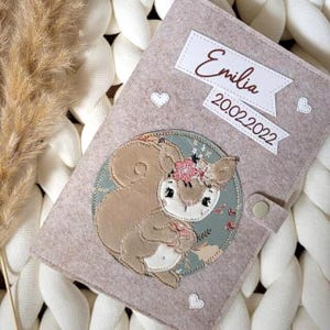Könnte beinhalten: Ein hellrosa Filz-Buchumschlag mit Knopfverschluss. Er zeigt ein gesticktes Eichhörnchen mit Blumenkrone, ein Banner mit dem Namen "Emilia" und dem Datum "20.02.2022". Kleine weiße Herzen sind über den Umschlag verteilt.