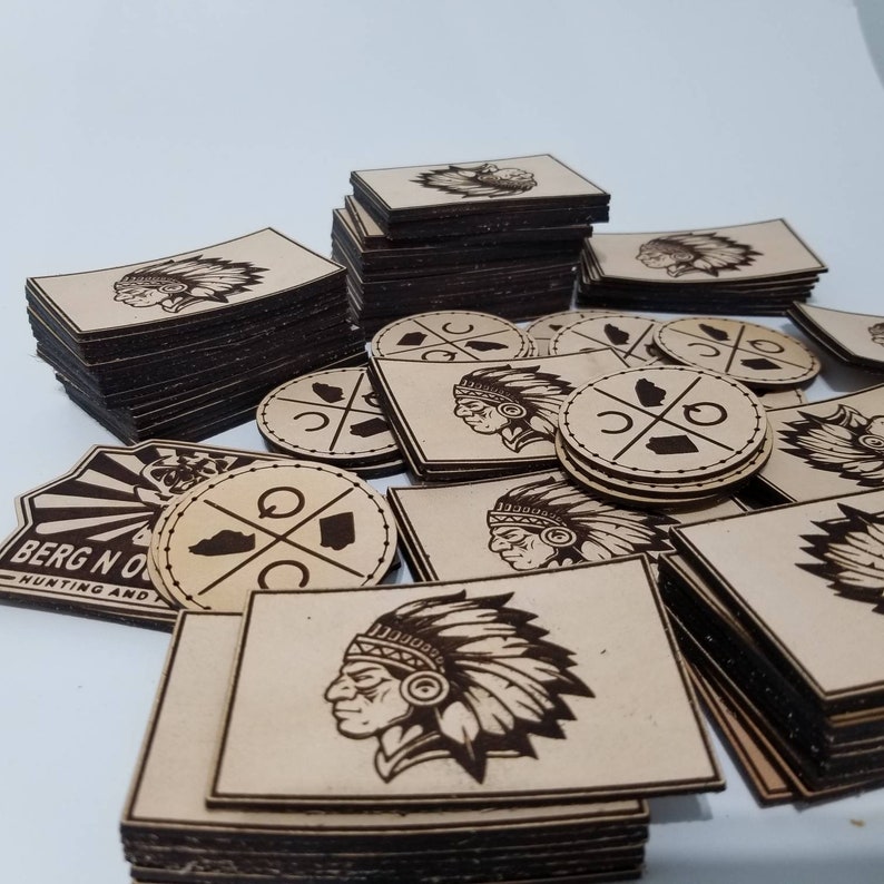 Puede incluir: Una pila de parches de madera cortados con l&aacute;ser que presentan un dise&ntilde;o de jefe nativo americano, un contorno de estado y un c&iacute;rculo con una cruz en su interior. Los parches son de varios tama&ntilde;os y est&aacute;n apilados sobre una superficie blanca.
