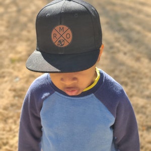 May include: A young person wearing a black baseball cap with a brown leather patch that says "MRO" with a globe inside the letters. The cap has a black brim and a blue and navy blue shirt.