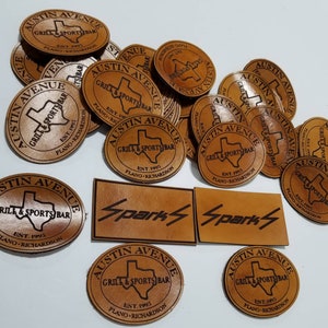 Puede incluir: Una colecci&oacute;n de parches de cuero marr&oacute;n con el texto "Austin Avenue Grill & Sports Bar Est. 1995 Plano-Richardson" y un gr&aacute;fico del estado de Texas. Dos parches adicionales dicen "Sparks" en una fuente estilizada.
