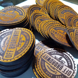 Può includere: Un mucchio di toppe rotonde in pelle marrone con il testo "Off Broadway" e "Taproom" in un font stilizzato. Le toppe hanno un aspetto vissuto e sono decorate con un piccolo grafico di una boccale di birra. Il testo "Madison, Indiana" e "Fondato nel 2014" è incluso anche sulle toppe.