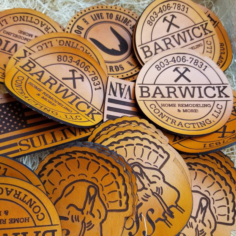 Puede incluir: Primer plano de varios parches de cuero con el texto "Barwick" y un n&uacute;mero de tel&eacute;fono. Los parches son marrones y tienen un aspecto desgastado. El texto es negro y los parches est&aacute;n dispuestos en una pila.