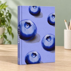Pode incluir: Um caderno de capa dura em roxo claro com um padrão repetitivo de mirtilos azul-escuro em estilo aguarela. A palavra Notebook está impressa verticalmente em texto branco ao longo do lado esquerdo da capa frontal.