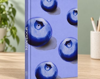 May include: A hardcover notebook with a light purple cover featuring a repeating pattern of dark blue watercolor-style blueberries. The word Notebook is printed vertically in white text along the left side of the front cover.