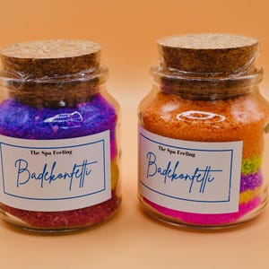 Może przedstawiać: Dwa szklane słoiki z korkowymi pokrywkami wypełnione kolorowymi konfetti do kąpieli. Słoiki mają białe etykiety z tekstem "The Spa Feeling Badekonfetti" nadrukowanym niebieskim kolorem.