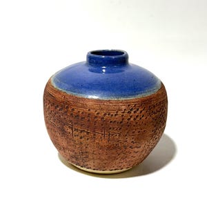 May include: A handcrafted ceramic vase with a rounded shape. The top is a vibrant blue, contrasting with the textured, reddish-brown body. The vase has a small opening and a unique, dotted pattern.