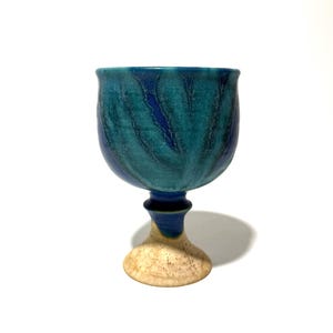 May include: A handmade ceramic goblet with a teal and blue glaze. The cup has a rounded bowl shape and a stem with a beige base. The goblet is approximately 6 inches tall.
