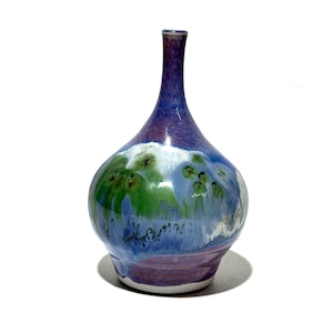 May include: A handcrafted ceramic vase with a bulbous body and a slender neck. The vase features a blend of colors, including shades of blue, green, and purple, with a white accent. The glaze creates a textured, artistic appearance, suitable for home decor.
