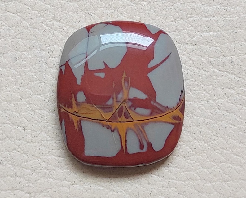 Pu&ograve; includere: Un cabochon di diaspro rosso, bianco e giallo con una superficie lucida e liscia. La pietra presenta un motivo unico e vorticoso.