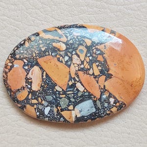 Puede incluir: Primer plano de una piedra ovalada pulida con un patr&oacute;n &uacute;nico de negro, marr&oacute;n y naranja. La piedra tiene un acabado liso y brillante y es perfecta para la elaboraci&oacute;n de joyas o manualidades.