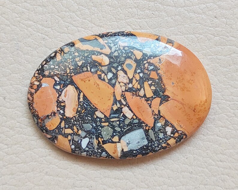 Puede incluir: Primer plano de una piedra ovalada pulida con un patr&oacute;n &uacute;nico de negro, marr&oacute;n y naranja. La piedra tiene un acabado liso y brillante y es perfecta para la elaboraci&oacute;n de joyas o manualidades.