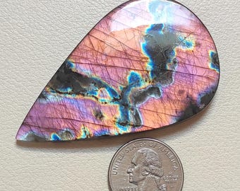 May include: A teardrop-shaped labradorite gemstone featuring iridescent pink, purple, and blue flashes with dark inclusions and fine surface fractures, positioned next to a United States quarter coin for scale to show its relative size.