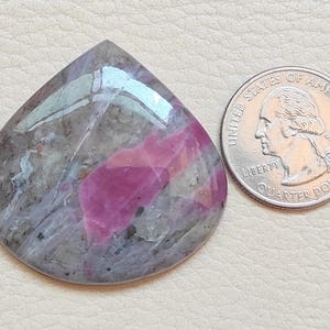 May include: A pink and grey cabochon gemstone with a teardrop shape. The gemstone is about the size of a UK 20 pence coin.