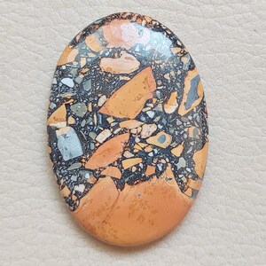 Puede incluir: Primer plano de una piedra lisa de forma ovalada con un patr&oacute;n &uacute;nico de negro, naranja y blanco. La piedra tiene un acabado pulido y es perfecta para la elaboraci&oacute;n de joyas o manualidades.