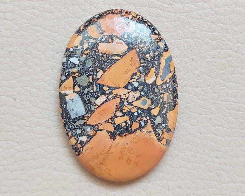 Puede incluir: Primer plano de una piedra lisa de forma ovalada con un patr&oacute;n &uacute;nico de negro, naranja y blanco. La piedra tiene un acabado pulido y es perfecta para la elaboraci&oacute;n de joyas o manualidades.