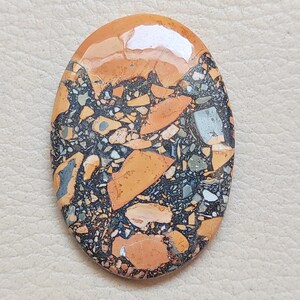 Puede incluir: Un cabuj&oacute;n de jaspe pulido, naranja, marr&oacute;n y negro con un patr&oacute;n de remolino. La piedra tiene forma ovalada y una superficie lisa y pulida.