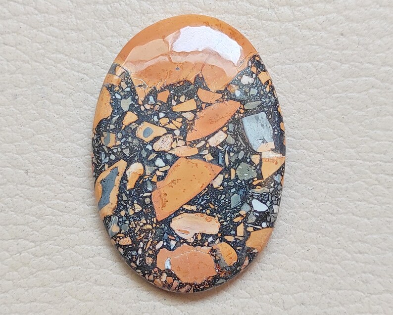 Puede incluir: Un cabuj&oacute;n de jaspe pulido, naranja, marr&oacute;n y negro con un patr&oacute;n de remolino. La piedra tiene forma ovalada y una superficie lisa y pulida.