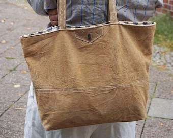 May include: A large, tan tote bag with sturdy straps is carried over the shoulder. The bag has a textured appearance and a small pocket on the front. The interior lining features a black and white checkered pattern. The bag is made of a durable material.