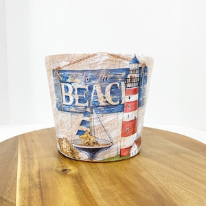 May include: A white ceramic planter with a nautical theme. The planter features a lighthouse, a sailboat, and a sign that says "to the beach".